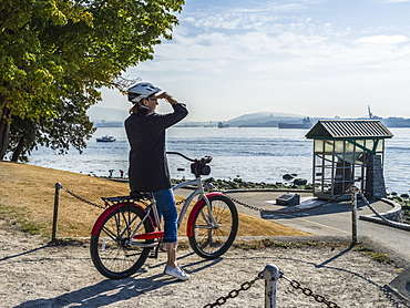 A woman sits on a bike looking out to the ocean and coastline at the Stanley Park Seawall, Vancouver, British Columbia, Canada