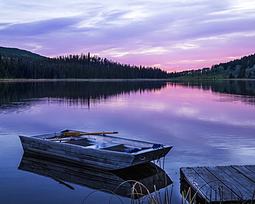 A wooden rowboat sits beside a dock on a tranquil lake reflecting the pink of a sunset, Lac Le Jeune Provincial Park, Kamloops, British Columbia, Canada