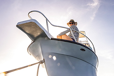 A woman wearing sunglasses and a sun hat looks down at the camera from the deck of a boat against a blue sky with sunlight, Bay Islands Department, Honduras