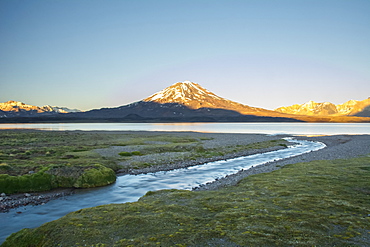 A stream leads the eye towards a high altitude lake. A volcano and snow-capped mountains in the distance are lit by the sunrise against a pure blue sky, Mendoza, Argentina