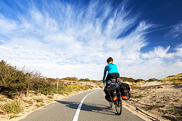 Female cyclist on paved bike pathway along rolling sand dunes with dramatic clouds and blue sky, South of Zandvoort, Netherlands