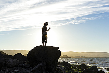 A woman stands on a rock looking out along the coast at sunset, silhouetted and backlit by the sunlight, San Mateo, California, United States of America