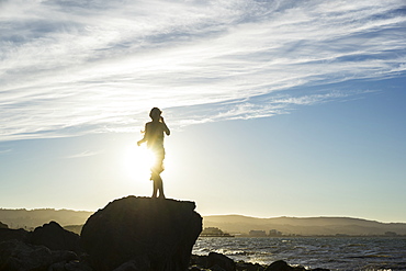 A woman stands on a rock looking out along the coast at sunset, silhouetted and backlit by the sunlight, San Mateo, California, United States of America