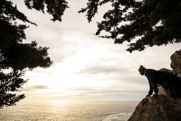 A man looks down over a rock to the ocean below at dusk, Julia Pfeiffer Burns State Park, California, United States of America