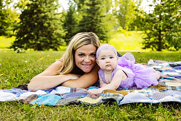 A mother and her baby daughter laying on a picnic blanket and posing for the camera while enjoying a family outing in a city park on a warm fall afternoon, Edmonton, Alberta, Canada