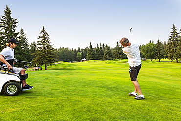 A male golfer drives a golf ball down the green with a wedge on a golf course while a disabled golfer in a specialized wheelchair watches, Edmonton, Alberta, Canada