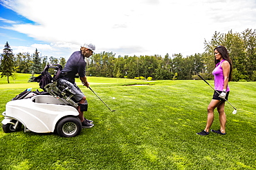 A physically disabled golfer, using a specialized wheelchair, hits the golf ball with his golf club on the golf green as a female golfer stands watching, Edmonton, Alberta, Canada