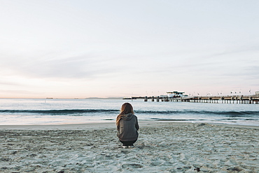 A girl crouches on the beach looking out over the ocean to the horizon at sunset, Long Beach, California, United States of America