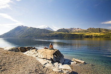 A woman basking in the sun on the shore of a lake with a snow-capped volcano in the distance, Neuquen, Argentina