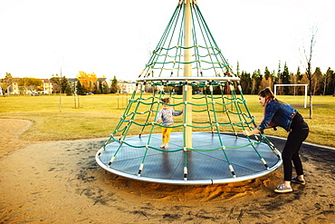 A young mom spinning her daughter while playing on a merry go round with a rope climber in a playground at sunset during a warm autumn evening, Edmonton, Alberta, Canada