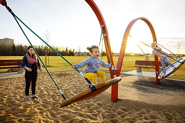 A young mom and her daughters playing on saucer swings in a playground on a warm autumn evening, Edmonton, Alberta, Canada