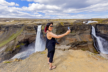 A young asian female hiker poses for a self-portrait on the edge of the Haifoss waterfall valley, a popular hiking destination in Iceland, Iceland