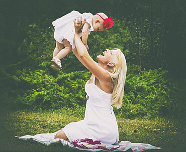 A beautiful young mother with long blonde hair enjoying quality time with her cute baby daughter and tossing her in the air while sitting on the grass in a city park during a summer day, Edmonton, Alberta, Canada