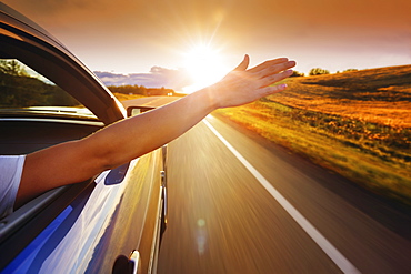 A woman's hand reaches out the window of a vehicle as it travels down the road into the sunset, Edmonton, Alberta, Canada