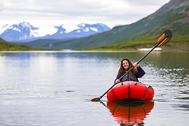 A woman paddles a packraft across Landmark Gap Lake, off the Denali highway, with the Alaska Range in the distance, Alaska, United States of America