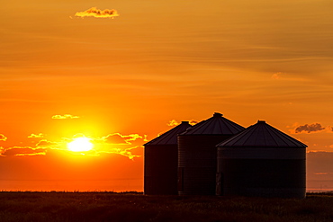 Silhouette of large metal grain bins at sunrise with orange sun rising over clouds, Alberta, Canada