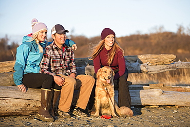 A young couple and a friend with a dog sit on a piece of driftwood on a beach looking out to the ocean at sunset, Anchorage, Alaska, United States of America