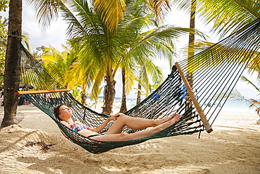 A young woman in a bikini lays in a hammock on a tropical beach with the ocean in the background, Negril, Jamaica