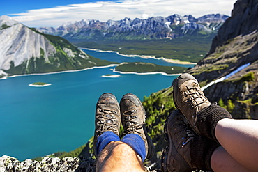 Close-up of a couple's hiking boots over a cliff overlooking a colourful alpine lake and mountain range in the distance, Kananaskis Country, Alberta, Canada