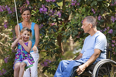 Man with spinal cord injury in wheelchair watching his daughter on swing along with wife