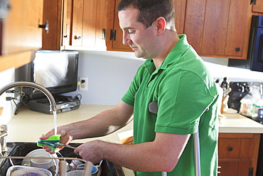 Man after anterior cruciate ligament (ACL) surgery with crutches washing dishes in the kitchen