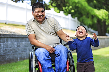 Hispanic man with Spinal Cord Injury in wheelchair with his son laughing in lawn