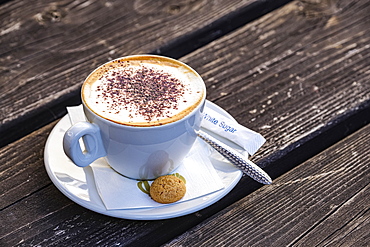 Cappuccino by an Italian cafe on wooden table, served with a small biscuit; Hexham, Northumberland, England