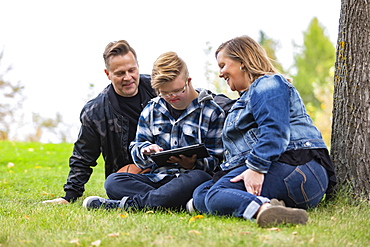 A young man with Down Syndrome learns a new program on a tablet with his father and mother while enjoying each other's company in a city park on a warm fall evening: Edmonton, Alberta, Canada