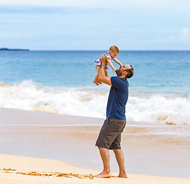 Father holds his baby daughter in the air as he stands on the beach next to the ocean; Maui, Hawaii, United States of America