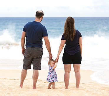 Mother and father hold the hands of their baby daughter as they stand looking out at the ocean; Maui, Hawaii, United States of America