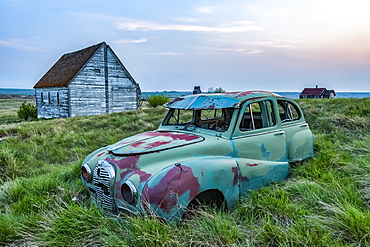 Vintage car sitting in the overgrown grass in a field with old buildings on a farmstead; Saskatchewan, Canada