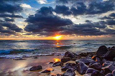 Sunrise over the Pacific Ocean from the rocks on the shore of Lydgate Beach and dark clouds overhead; Kapaa, Kauai, Hawaii, United States of America