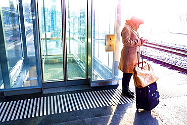 A woman stands with her suitcase on the platform of a train station beside the tracks and uses her smart phone; St. Gallen, St. Gallen, Switzerland