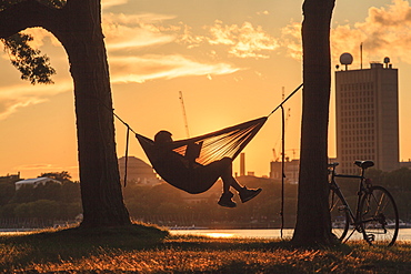 A young man sits reading in a hammock between two trees at the water's edge at sunset with a bicycle leaning against the tree and buildings across the water