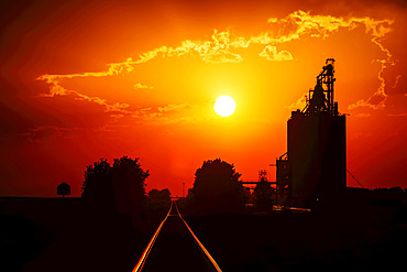 A grain storage facility at sunset with a bright yellow sun in the glowing red sky; Alberta, Canada