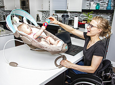 A young mom with a spinal cord injury looks after her newborn baby while working on her computer in the kitchen; Edmonton, Alberta, Canada