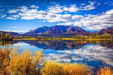 Mountain range reflects on Mentasta Lake with fall coloured foliage under blue sky, Tok cutoff from the Glenn Highway, South-central Alaska in autumn; Alaska, United States of America