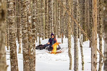 A woman with snowshoes sits on the snow and warms up by a campfire in a forest in wintertime, Gaspesie National Park; Quebec, Canada