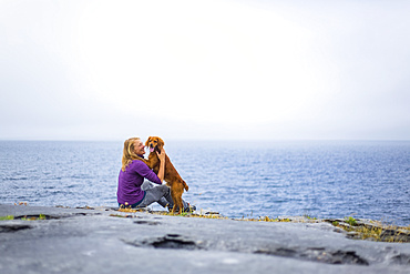 Woman embracing dog on a rocky ground overlooking the sea on a cloudy day; Fanore, County Clare, Ireland