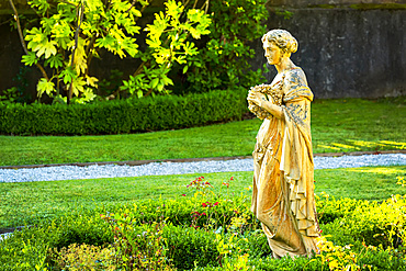 Stone female statue in garden with pebble pathway, framed by stone wall and trees; Cornwall County, England