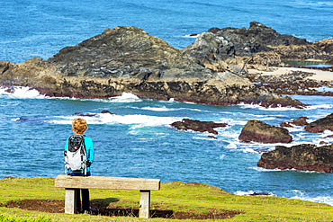 A female hiker sitting on a wooden bench overlooking rocky shoreline; Cornwall County, England