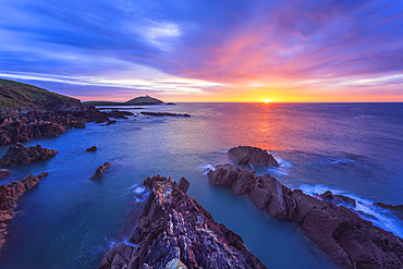 Sun rising over the Irish coastline with jagged rocks in the foreground and a lighthouse on an island in the distance; Ballycotton, County Cork, Ireland