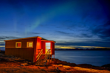 A small house on the water's edge with a view of the tranquil coastline at nightfall and the glowing northern lights above; Nuuk, Sermersooq, Greenland