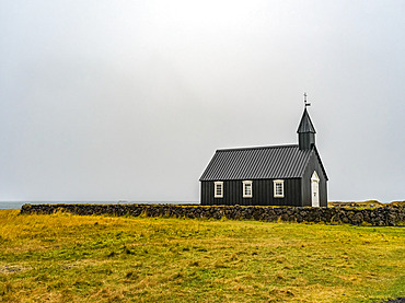 Church building with steeple and cross in a remote area with stone wall and grass; Snaefellsbaer, Western Region, Iceland