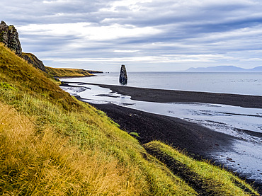 Tall rock formation and grassy slopes along the shoreline of a fjord; Iceland