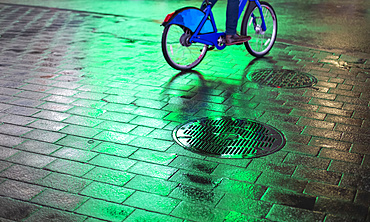 Cyclist riding bike on wet walkway with glowing green light at night in Manhattan; New York City, New York, United States of America