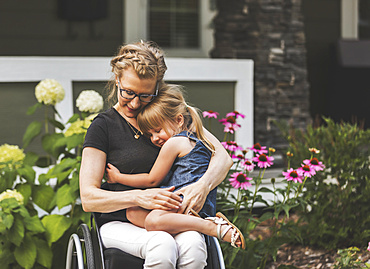 A paraplegic mom holding her little girl in her lap while sitting in her wheelchair in her front yard on a warm summer afternoon: Edmonton, Alberta, Canada.