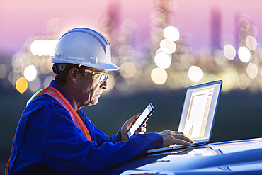 Man working on a laptop and smart phone with an oil refinery in the background; Alberta, Canada