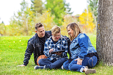 A young man with Down Syndrome learns a new program on a tablet with his father and mother while enjoying each other's company in a city park on a warm fall evening; Edmonton, Alberta, Canada