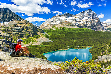 Male hiker sitting on rocky ridge overlooking blue alpine lake and mountains in the distance with blue sky and clouds, Yoho National Park; Field, British Columbia, Canada